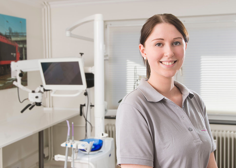 Eine Frau mit Pferdeschwanz steht in einem Behandlungsraum und lächelt in die Kamera